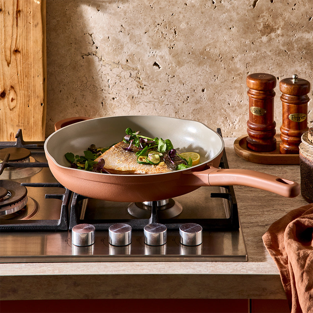 A nonstick frying pan with a beige interior sits on a gas stovetop, cooking a piece of fish with greens and lemon slices. The background shows a rustic stone wall, wooden salt and pepper shakers, and a textured kitchen setting.