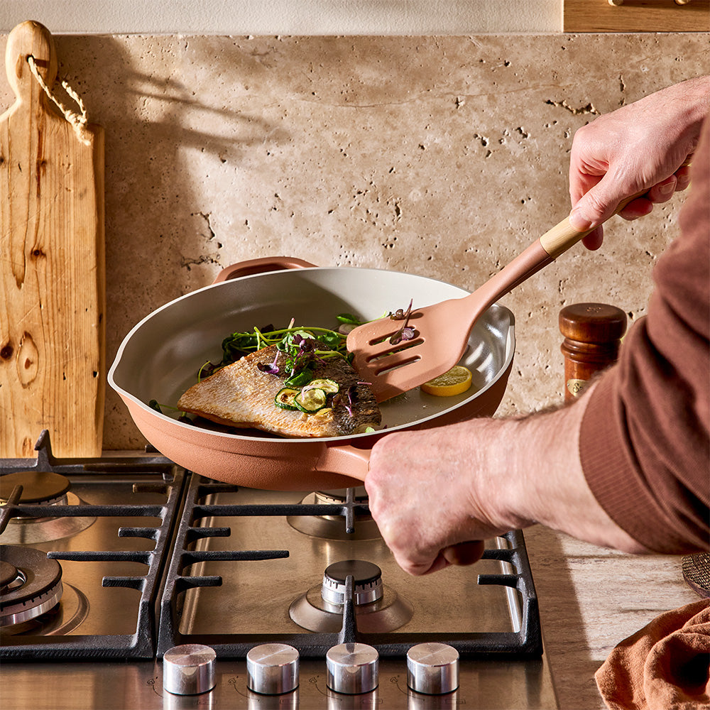 A person cooks food in a light beige non-stick frying pan with a dusty rose exterior, placed on a gas stove. A wooden spatula rests inside the pan, and a textured stone backsplash is visible in the background.