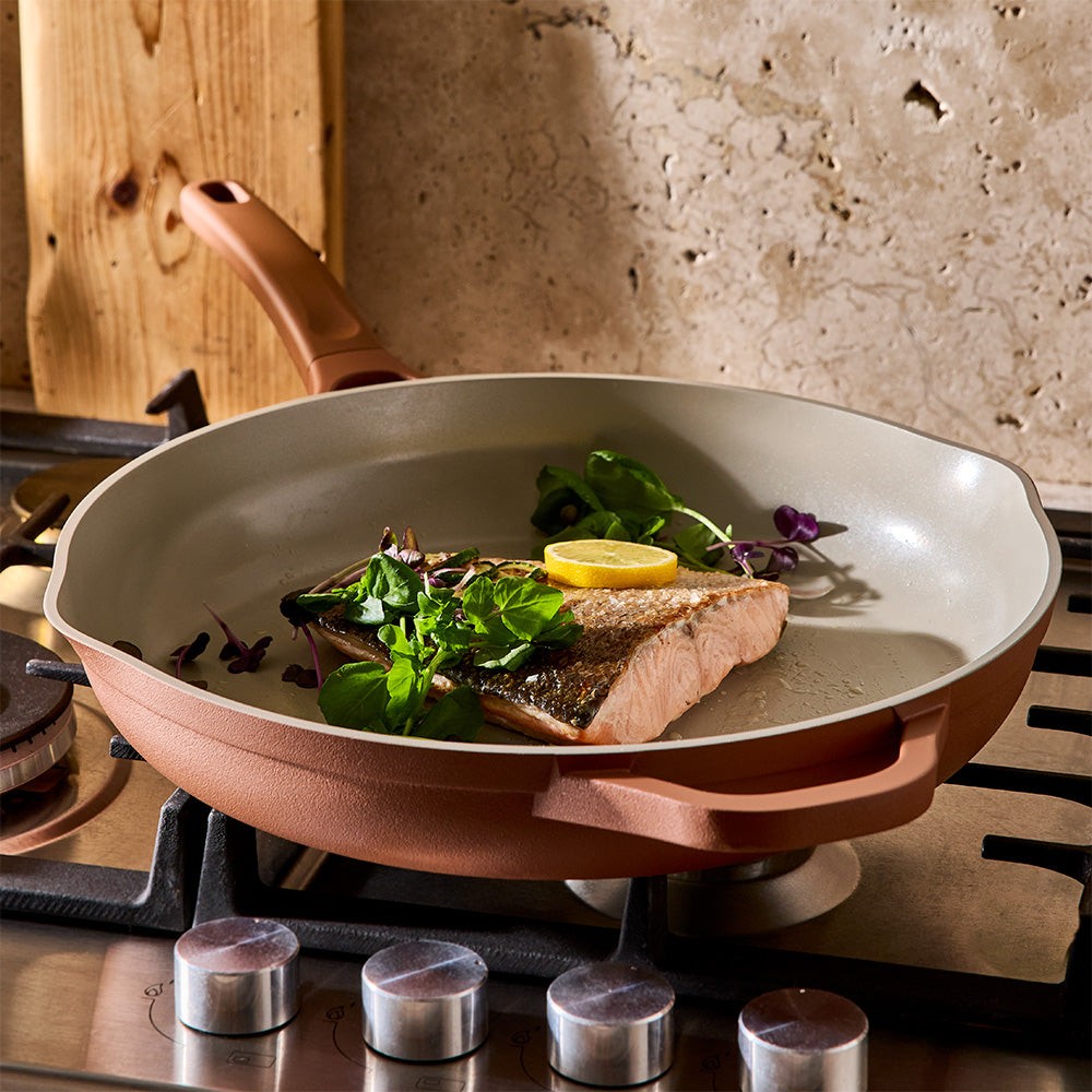 A non-stick frying pan with a copper-colored exterior and a light beige interior, shown on a gas stove with salmon cooking inside along with greens and a lemon slice.