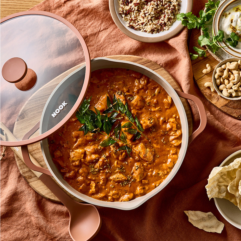 An overhead view shows cooked curry inside a terracotta-colored pan with its glass lid resting to the side on a peach tablecloth.