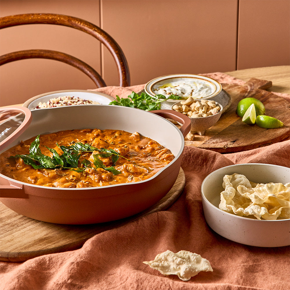 An uncovered terracotta-colored pan filled with cooked curry is placed on a table alongside side dishes.