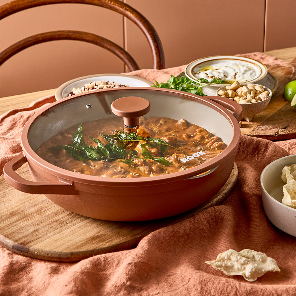 A terracotta-colored pan covered with a clear glass lid is filled with cooked curry on a dining table.