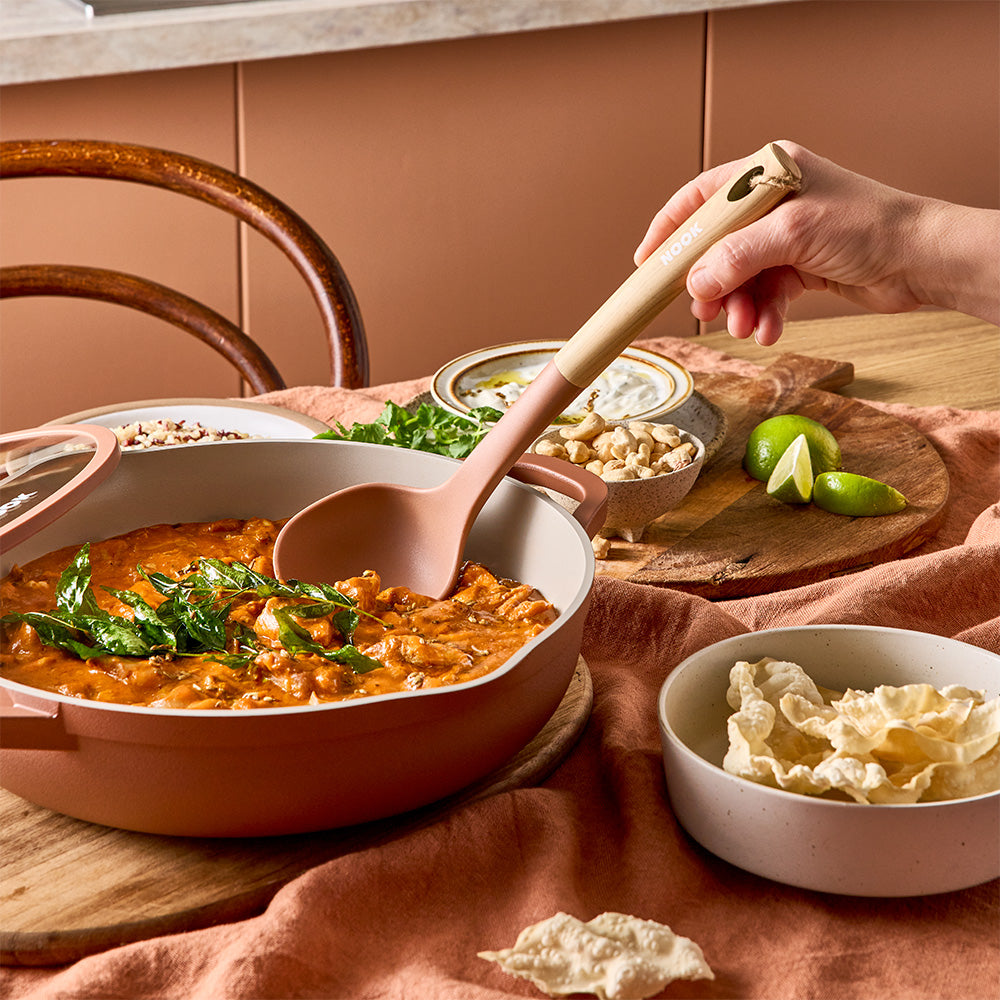 A person is using a wooden spoon to serve curry from a terracotta-colored pan set on a table with a peach tablecloth.