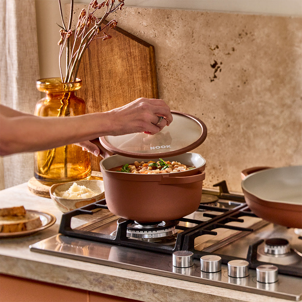 A person is using a hand to lift the glass lid off a terracotta-colored saucepan on a stovetop.