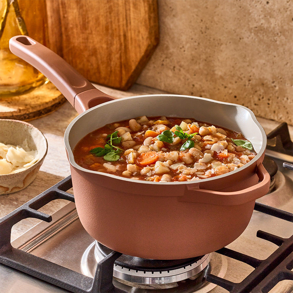 A terracotta-colored saucepan filled with bean soup is simmering on a gas stovetop burner.