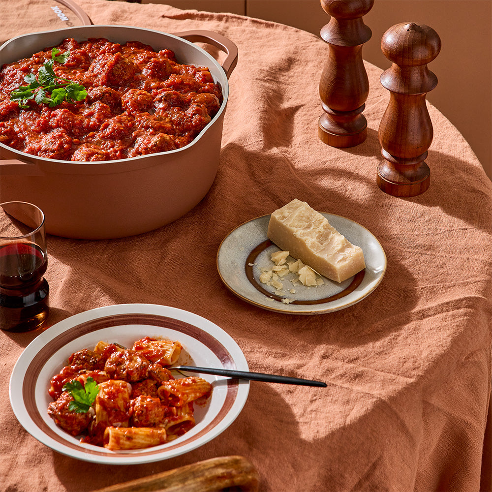 A terracotta-colored pot filled with meatballs is set on a table covered with a brown tablecloth next to cheese and wine.