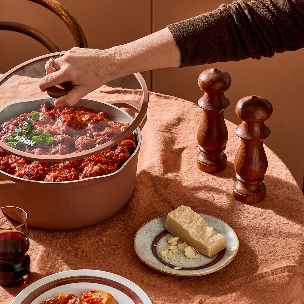 A person is using a hand to lift the glass lid off a terracotta-colored pot filled with meatballs.