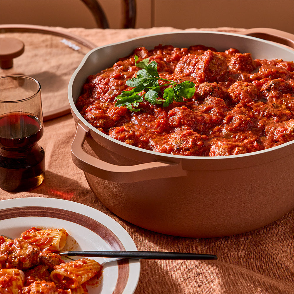 An uncovered terracotta-colored pot filled with meatballs in tomato sauce is placed on a table next to a served plate.