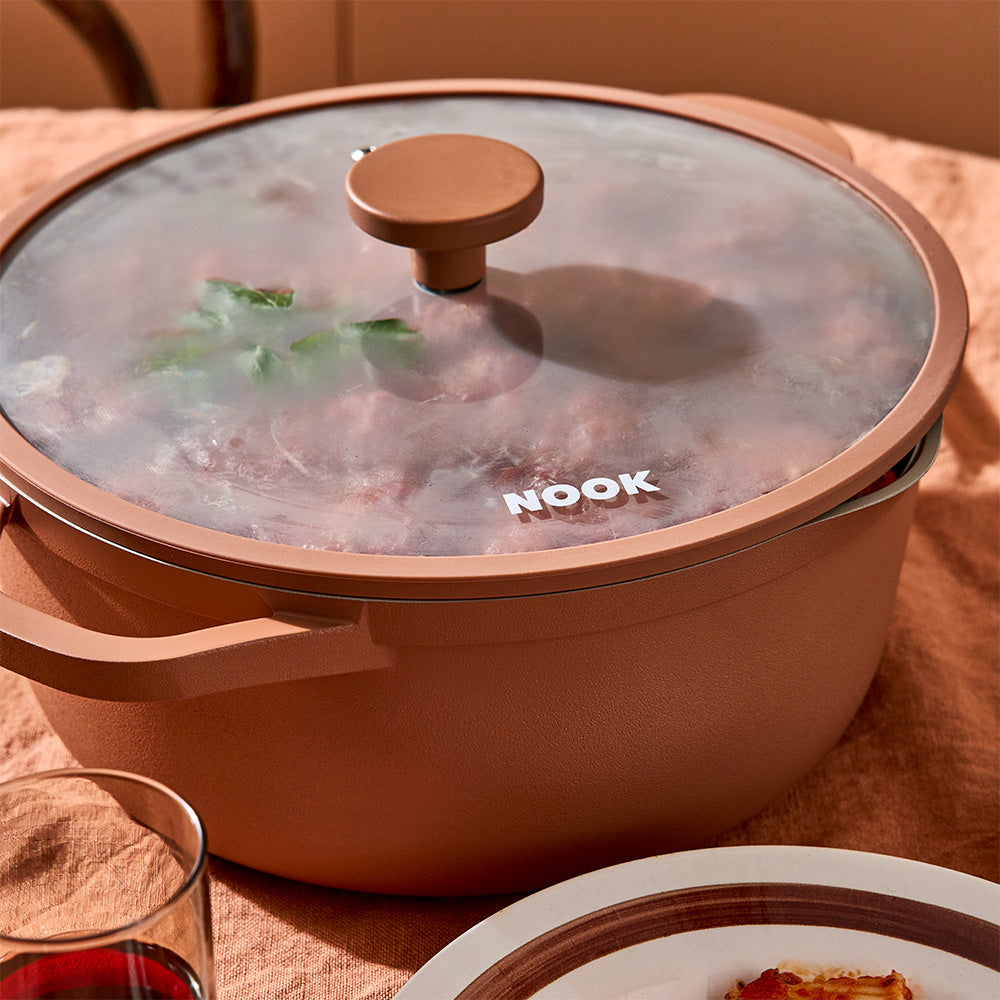 A close-up view focuses on a terracotta-colored pot filled with meatballs in tomato sauce and covered with a glass lid.