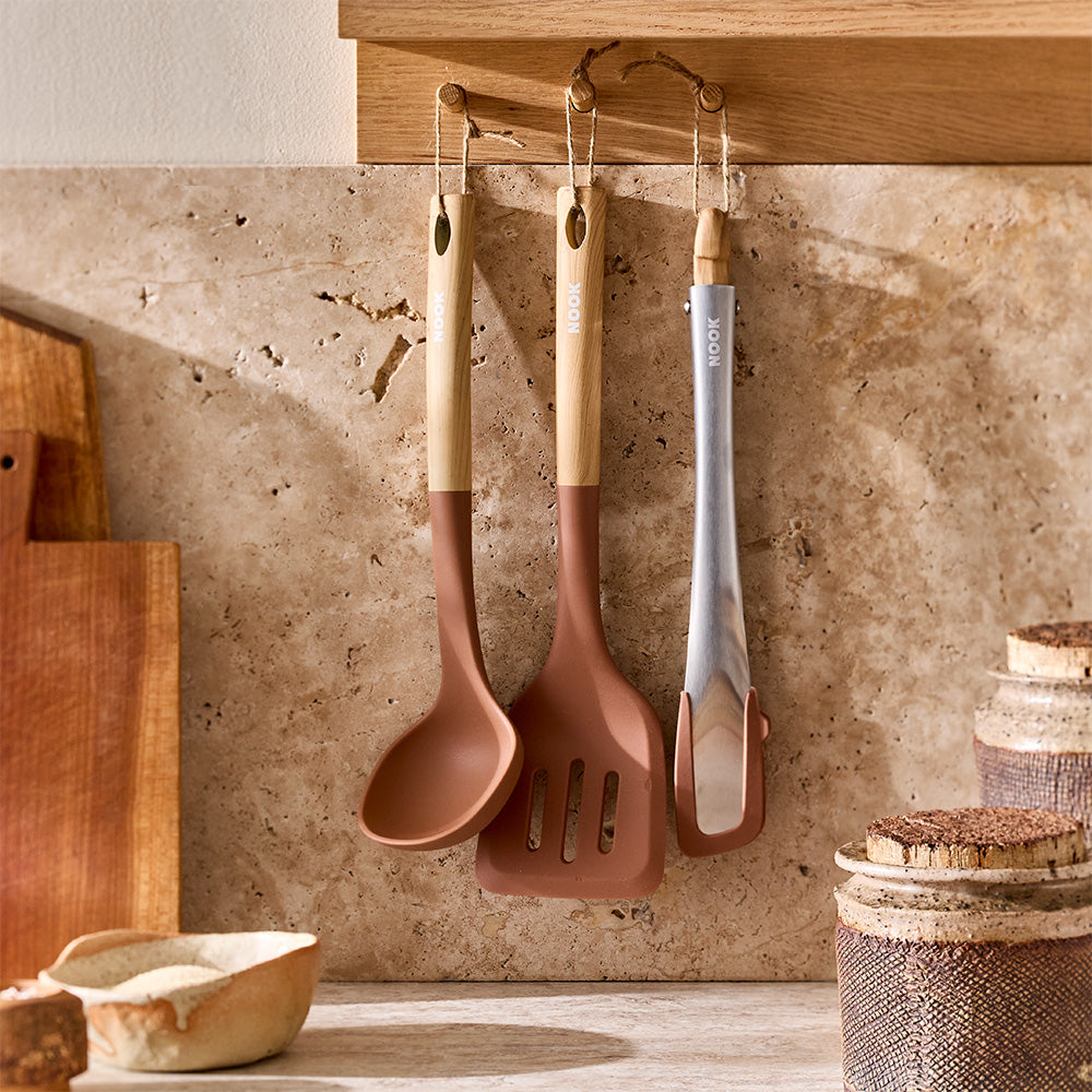 Brown silicone kitchen utensils with wooden handles are hanging on hooks against a warm tiled wall.