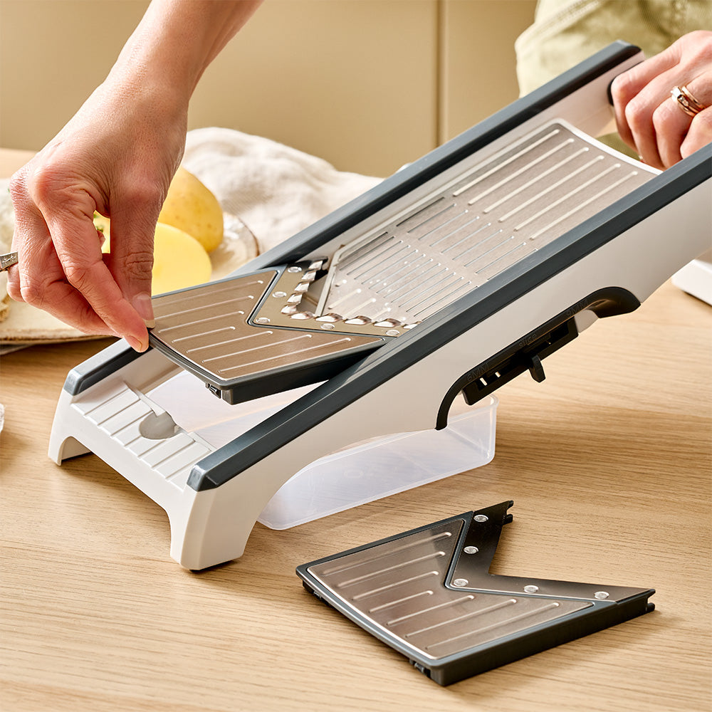 Hands are inserting an interchangeable cutting blade into the side track of the beige mandoline slicer resting on a wooden table.