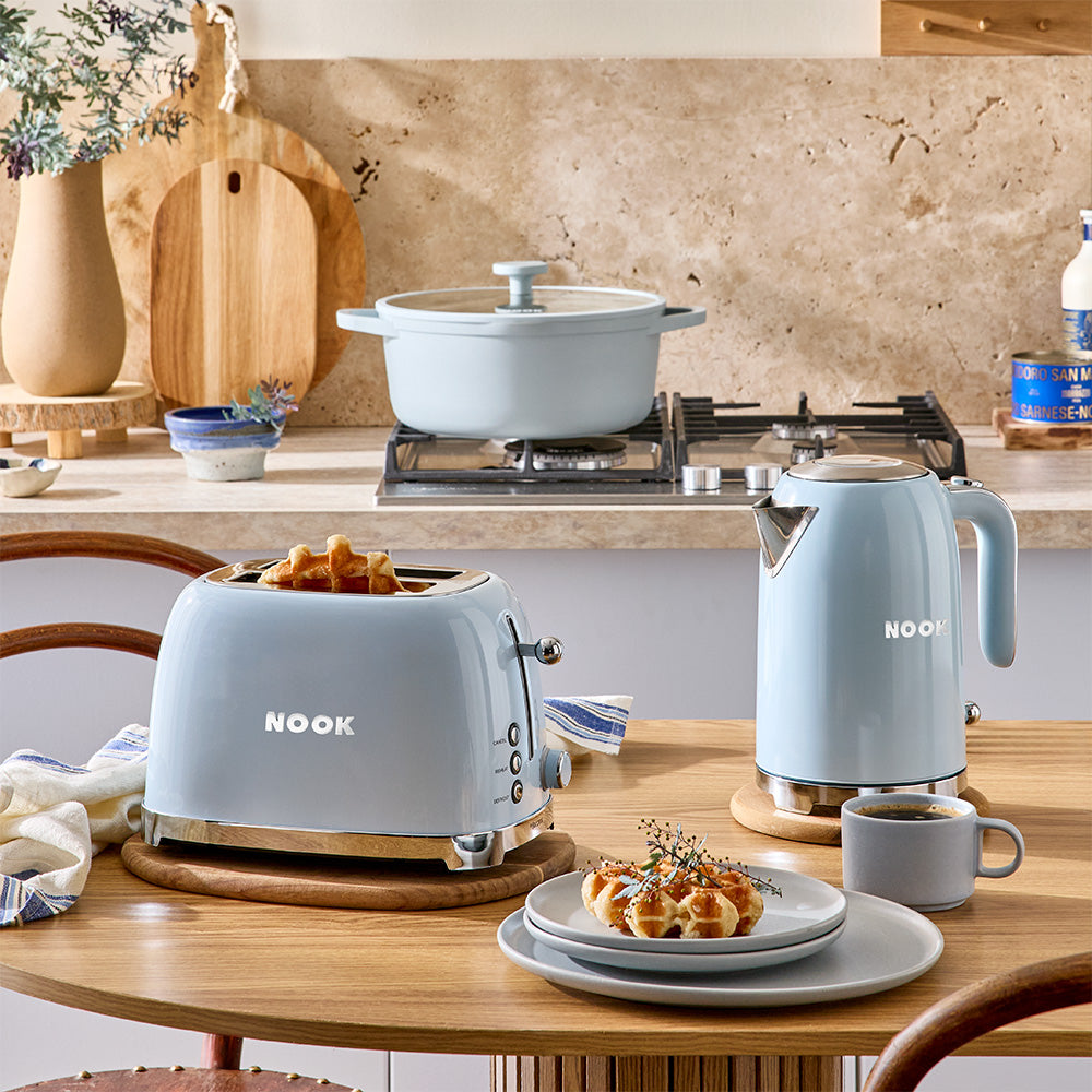 A light blue Nook toaster, kettle, and matching pot arranged on a wooden dining table in a warm kitchen setting, with food and a cup of coffee nearby.