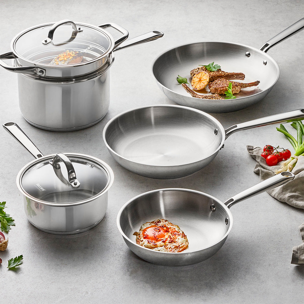 A set of stainless steel cookware including two saucepans with glass lids, a deep sauté pan, and two frying pans, arranged on a gray surface with food props.