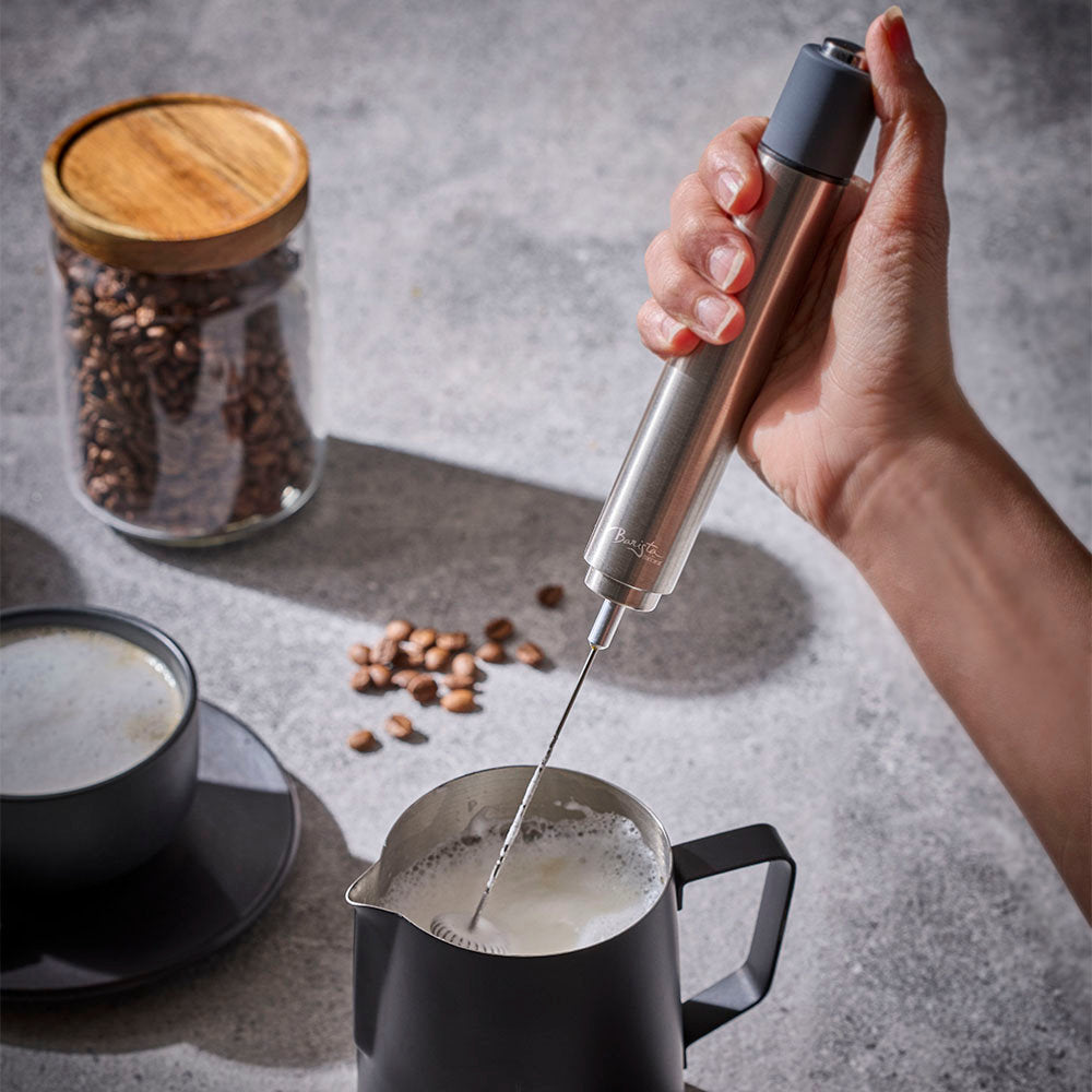 A hand holds a sleek stainless steel handheld milk frother over a black milk pitcher, creating foam. The scene is set on a gray stone surface with a cup of coffee, scattered coffee beans, and a glass jar of coffee beans in the background.