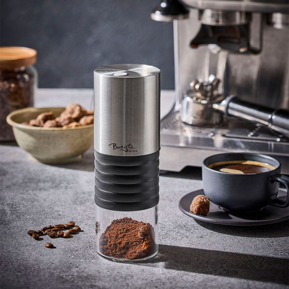 An electric coffee grinder is standing on a dark stone countertop next to an espresso machine, a cup of coffee, and coffee beans.