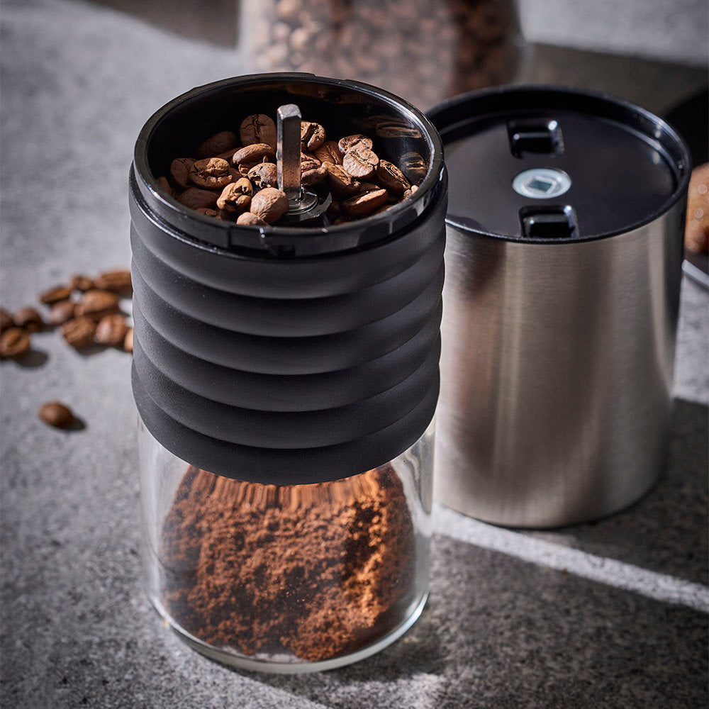 An electric coffee grinder with its top cap removed to reveal coffee beans inside the hopper is set on a dark textured surface next to its stainless steel lid.