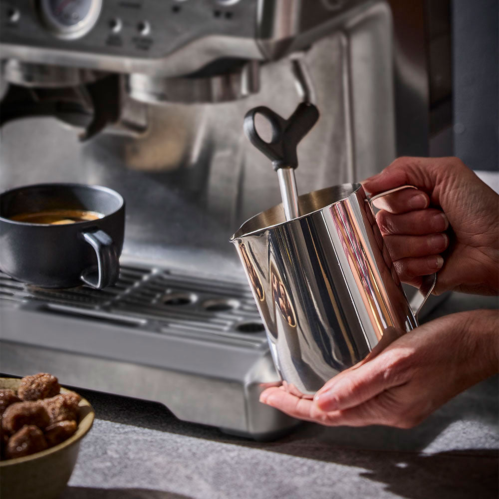 A stainless steel milk frother pitcher with a handle and steam wand, being held by hands in front of a stainless steel espresso machine on a countertop.