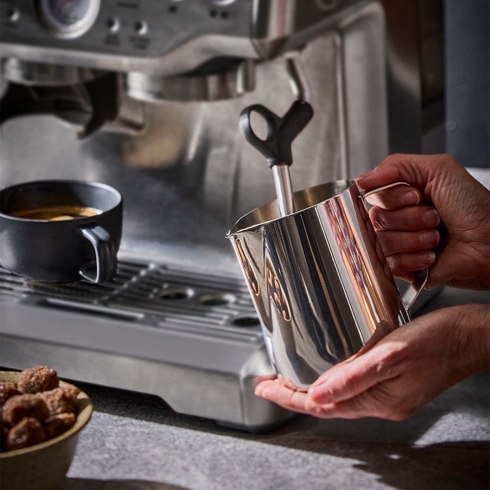 A stainless steel milk frother pitcher with a handle and steam wand, being held by hands in front of a stainless steel espresso machine on a countertop.