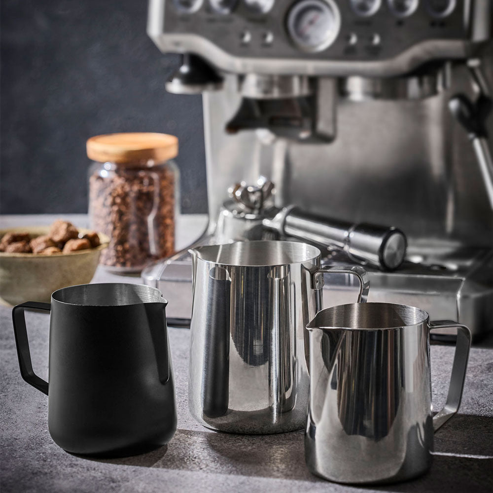 Three milk frothers in matte black and polished stainless steel arranged on a countertop in front of an espresso machine, with a jar of coffee beans and a bowl of nuts in the background.