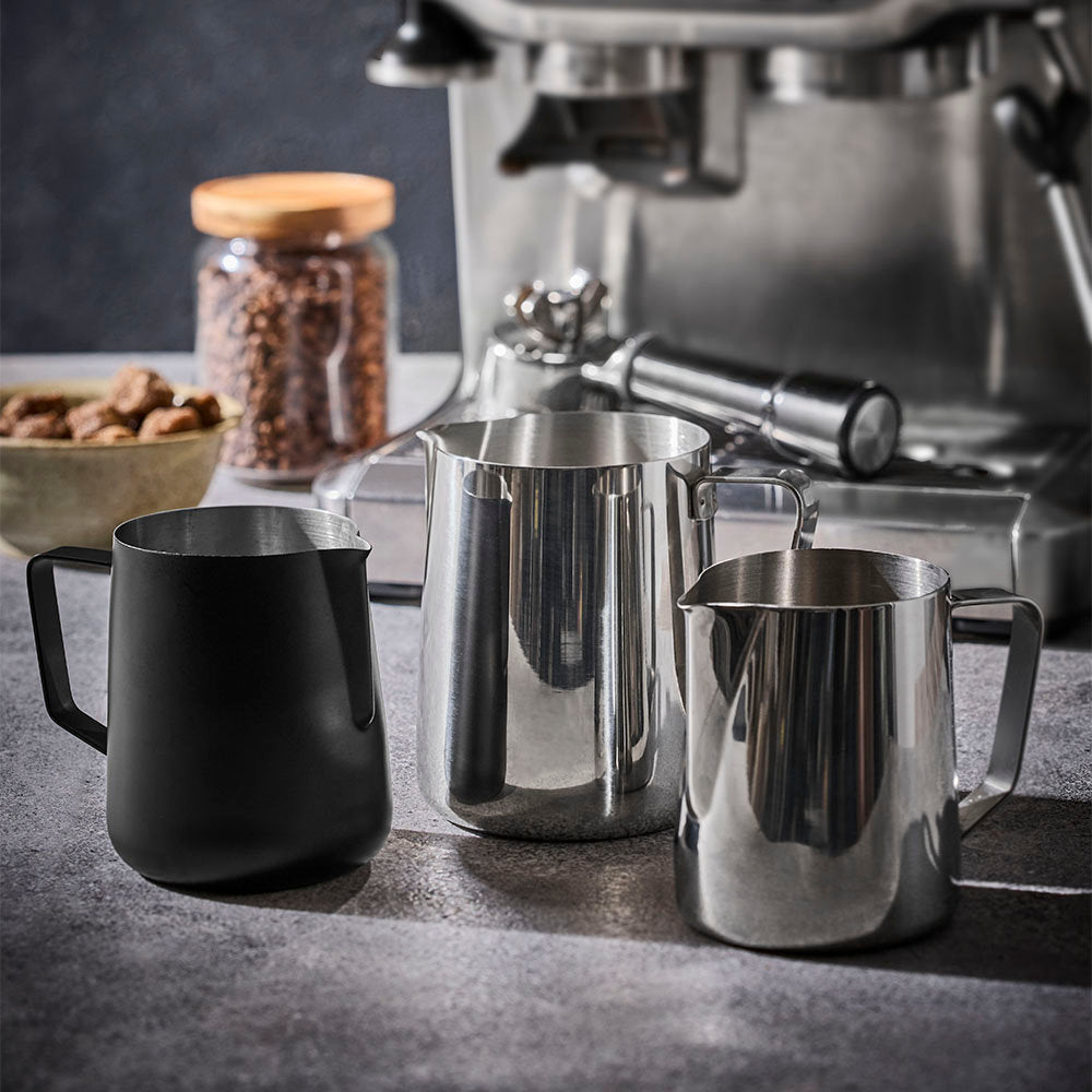 Three milk frothing pitchers in different finishes are arranged on a dark countertop in front of an espresso machine.