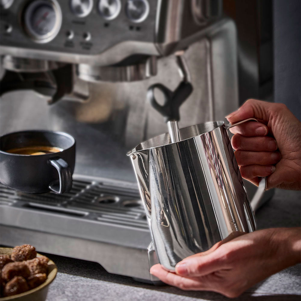 A close-up of a person's hands holding a shiny stainless steel milk pitcher under the steam wand of a professional espresso machine, with a cup of espresso and cookies on the counter in the background.
