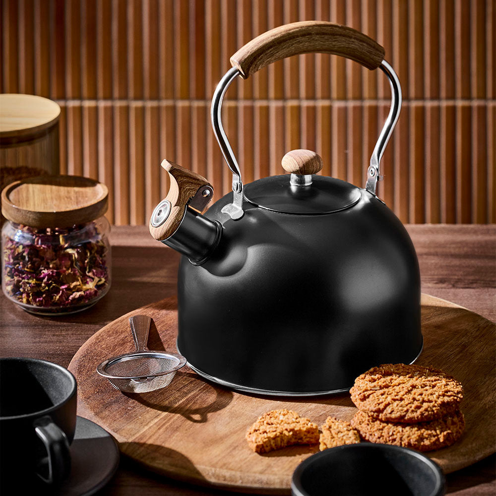 A black tea kettle is displayed on a wooden surface with tea snacks and spice jars in front of a textured wooden wall.