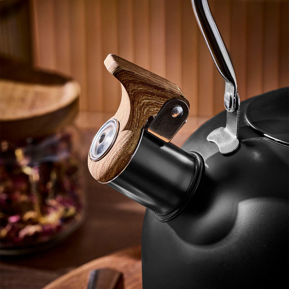 The spout of a black tea kettle is shown close up with its wooden cap closed, set against a blurred warm wooden background.