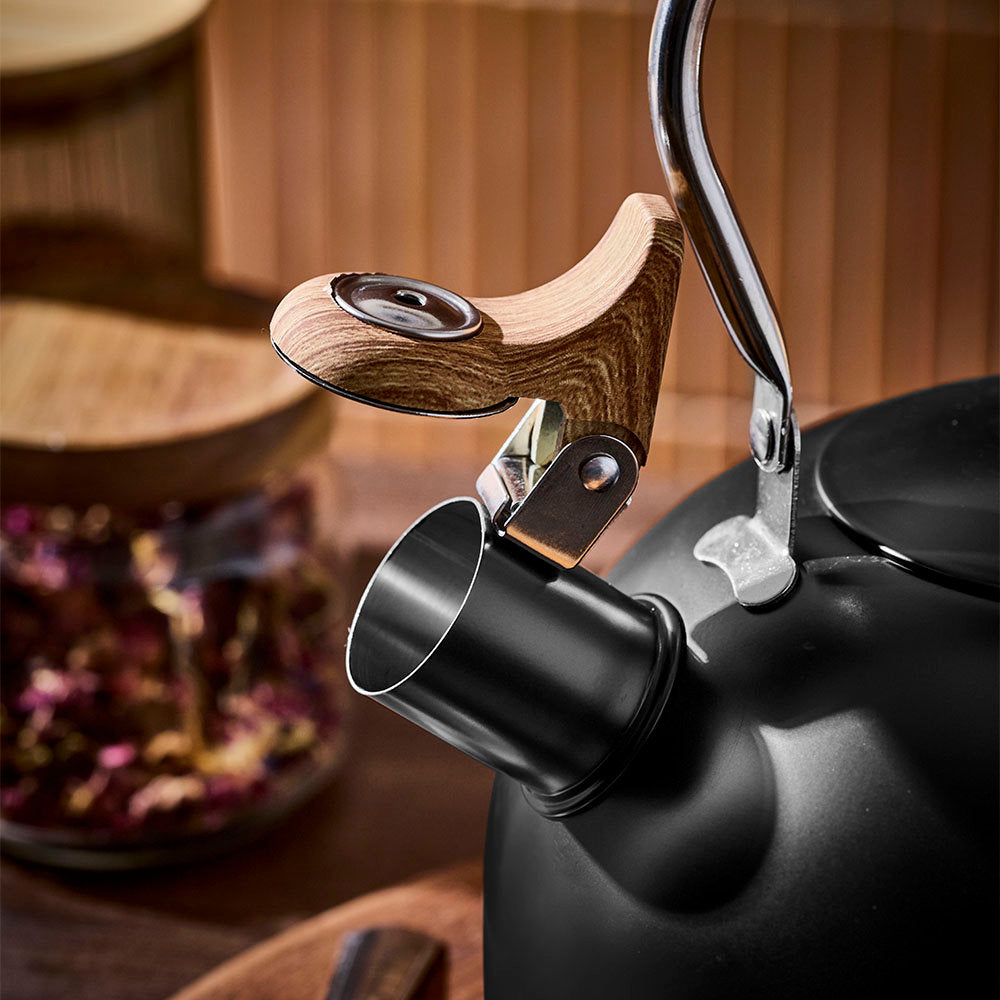 The spout of a black tea kettle is shown close up with its wooden cap open, set against a blurred warm wooden background.