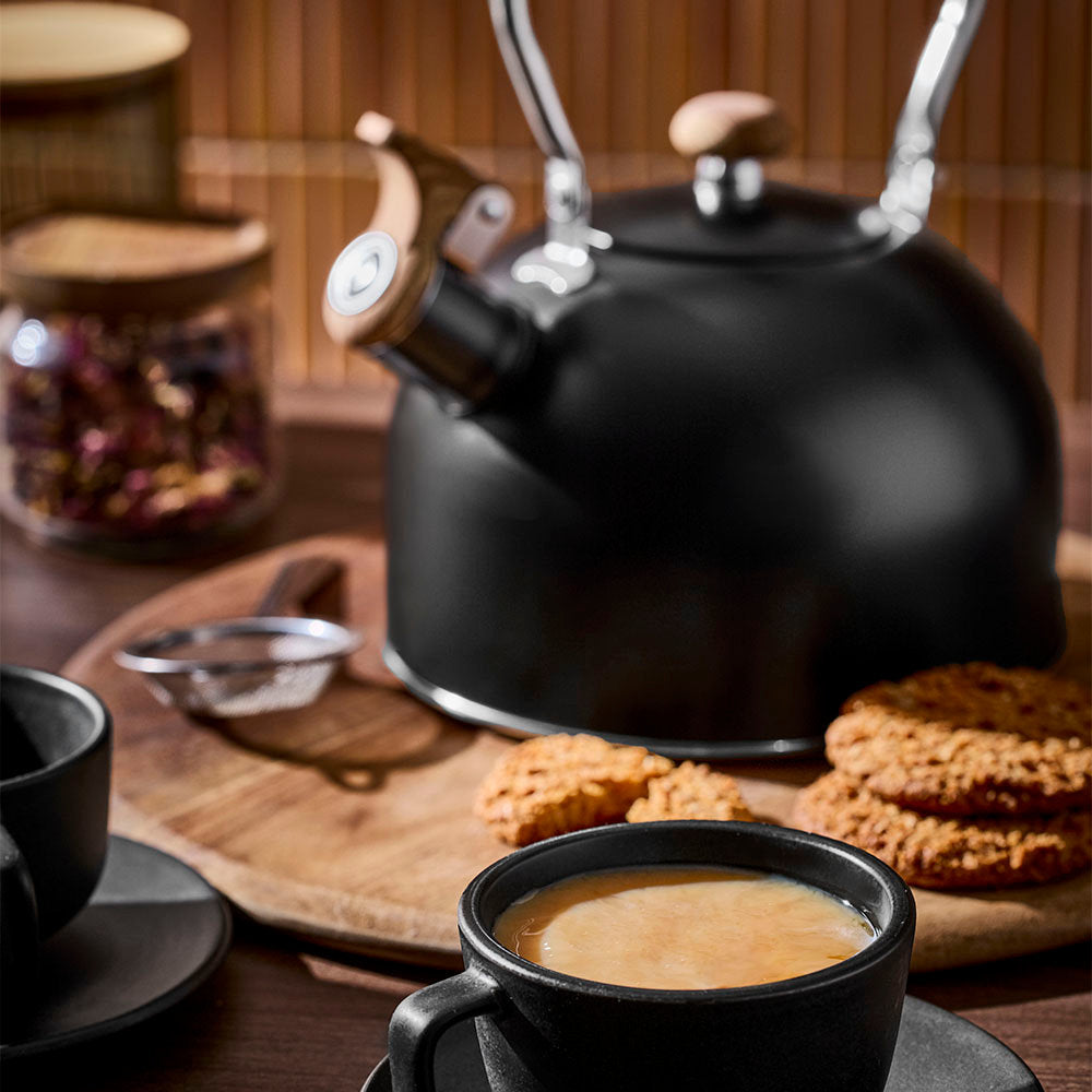 A black tea kettle is placed on a wooden board next to a coffee cup, cookies, and tea jars against a wooden slatted wall background.