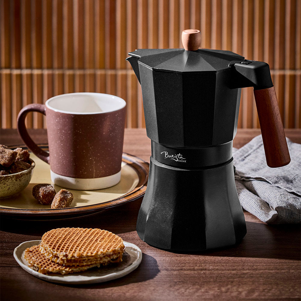 A matte black Moka pot with wooden handles is set on a dark wooden table beside a ceramic mug, waffles, and treats against a fluted wooden wall.