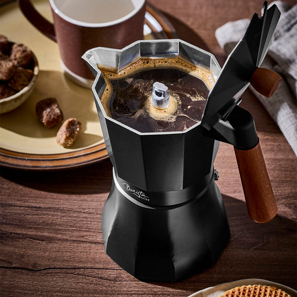 Hot brewed coffee is visible inside an open black Moka pot resting on a wooden surface with a cozy kitchen backdrop.