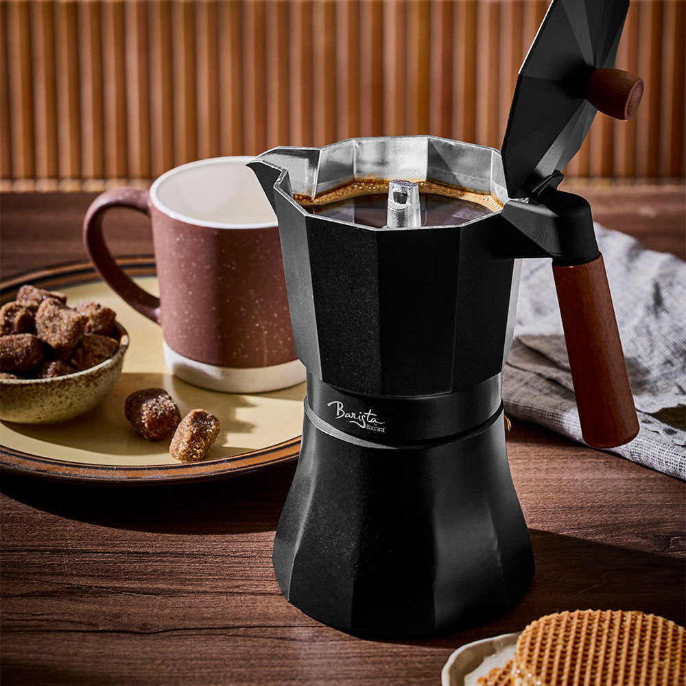 A black Moka pot with its lid open is placed on a wooden table alongside a mug and snacks against a slatted wooden background.