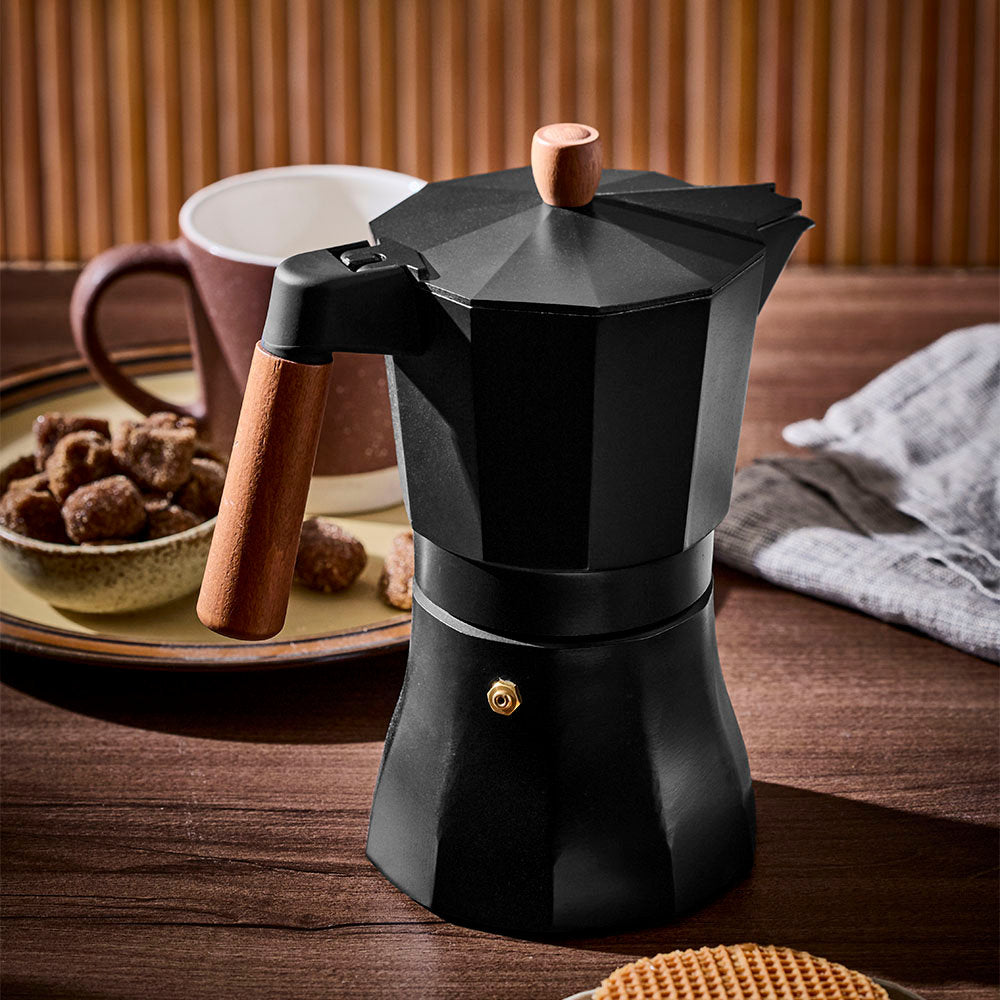 A closed black Moka pot is positioned prominently on a dark wooden tabletop, backed by a mug, snacks, and a textured wall.