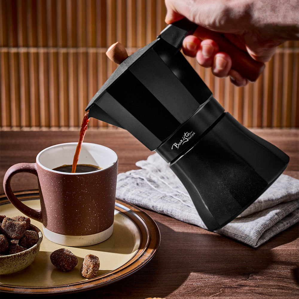 Fresh coffee is poured from a black Moka pot into a brown mug on a dark wooden surface with warm background paneling.