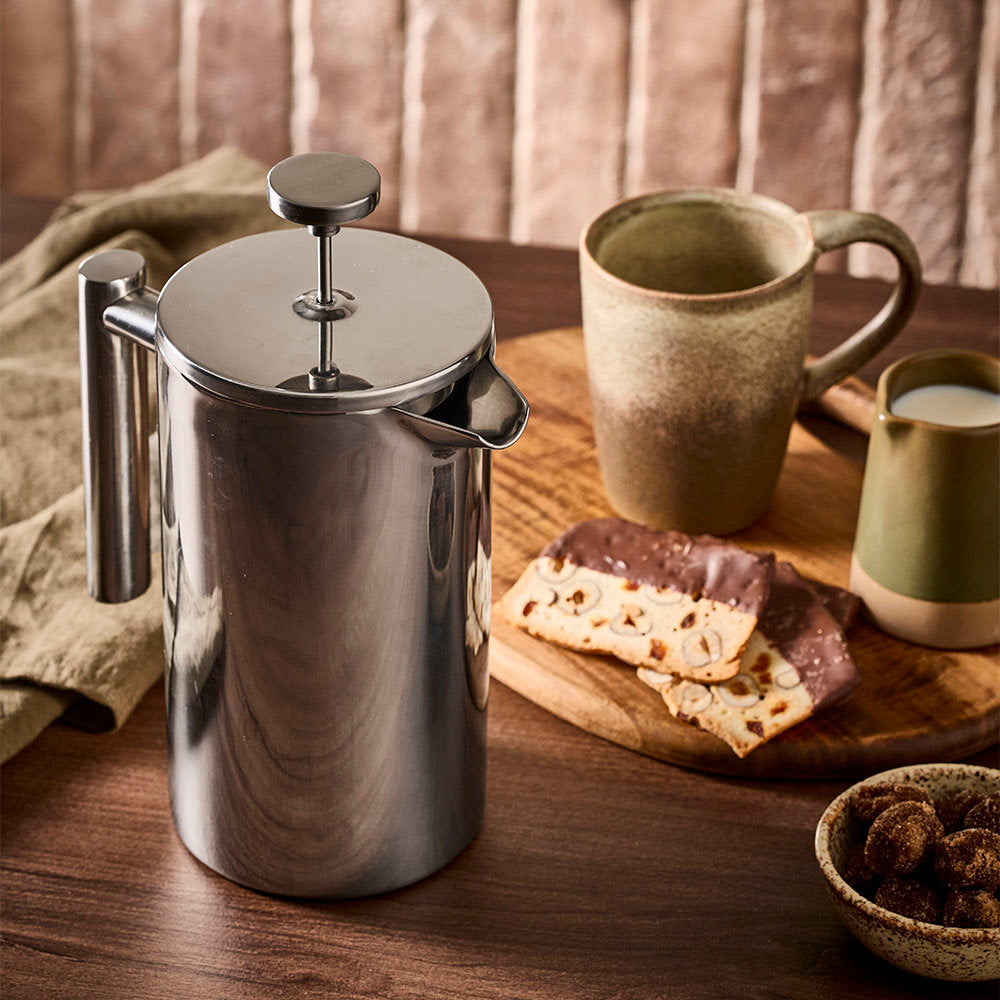 A stainless steel French press coffee maker sits on a wooden table in a warm kitchen setting, styled with ceramic mugs, biscotti, and a small bowl of chocolate-covered nuts.