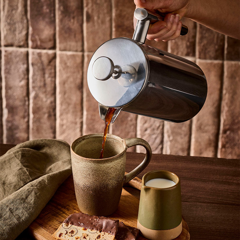 Fresh coffee is poured from a stainless steel French press into a ceramic mug on a wooden counter with a warm tiled backdrop.