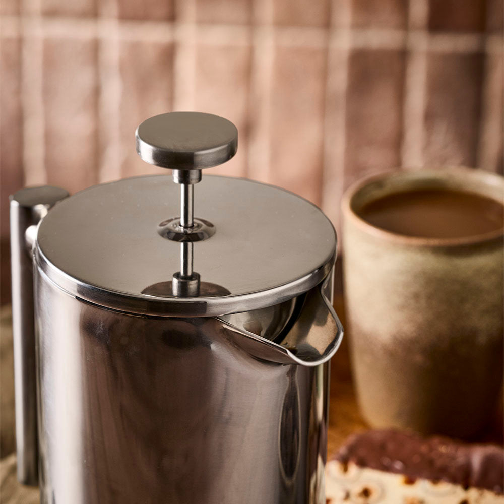 A close-up view of the stainless steel lid knob is highlighted on the French press against a cozy kitchen setting.