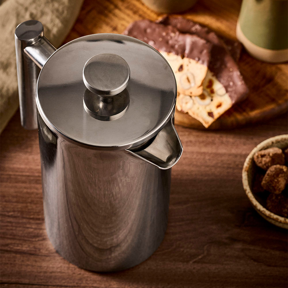 A top-angle view of a stainless steel French press is captured on a dark wooden counter with snacks in the background.