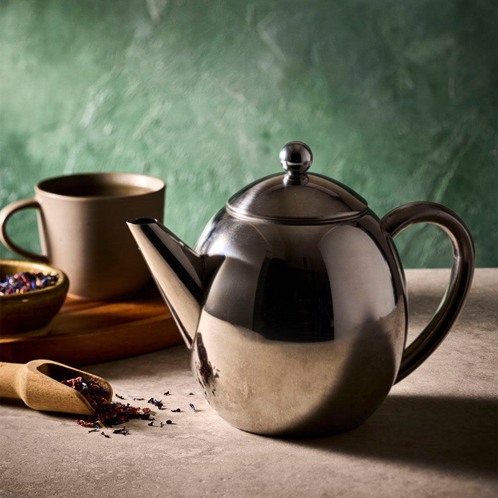A polished silver teapot with a matching lid sits on a tabletop next to a beige ceramic mug and a small bowl of loose tea, against a textured green background.