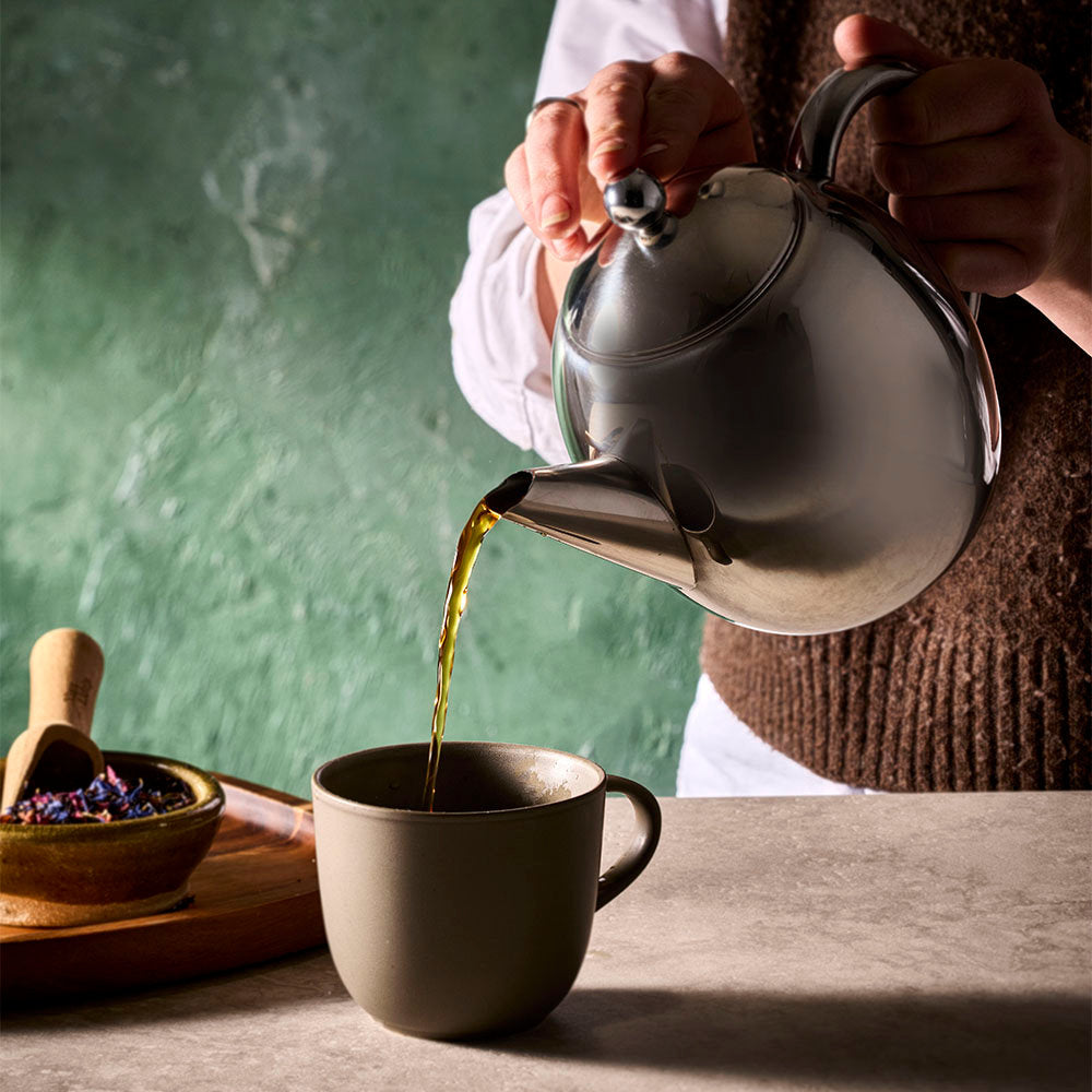 A person pours tea from a polished silver metal teapot into a matte dark gray ceramic cup, set against a textured green background.