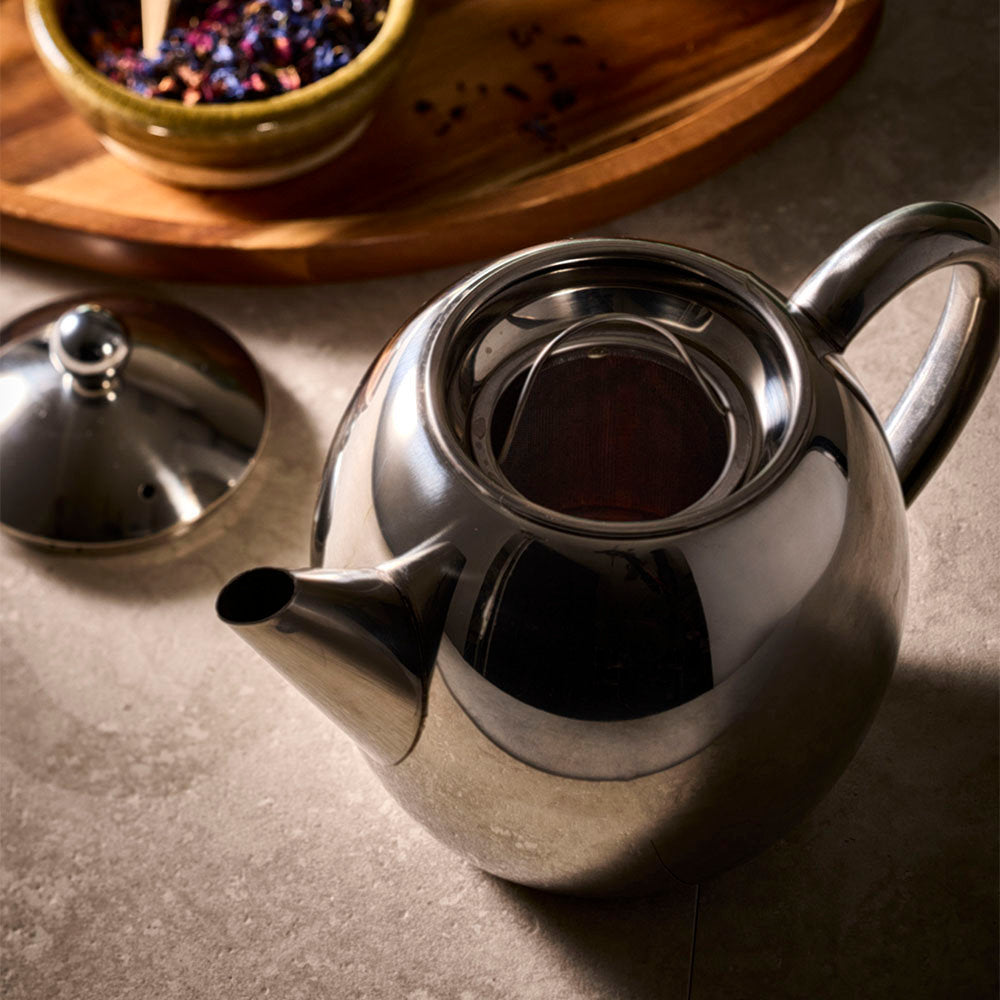 A polished stainless steel teapot with its lid off, placed on a stone-like surface. A small bowl of loose tea is visible in the blurred background on a wooden tray.