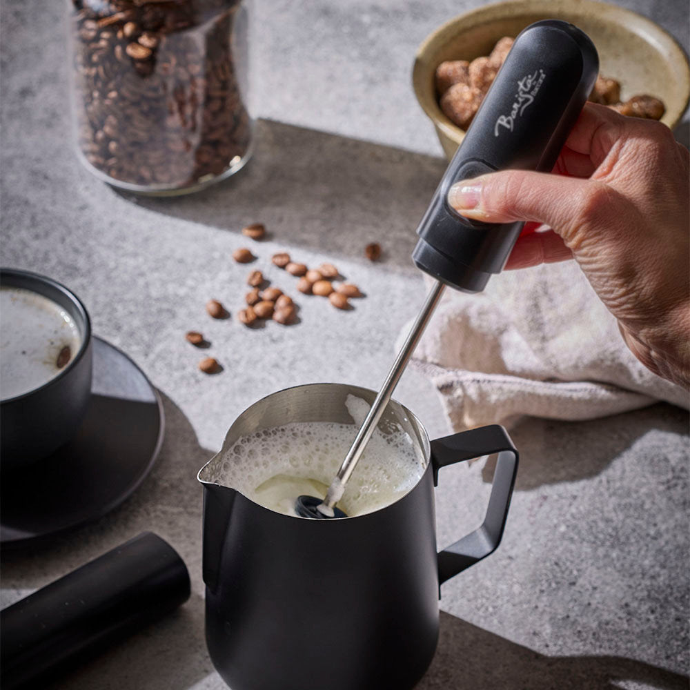 A hand holds a black handheld milk frother over a matte black milk pitcher, creating foam. The scene is styled with coffee beans, a cup of coffee, and a bowl of nuts on a gray textured surface.