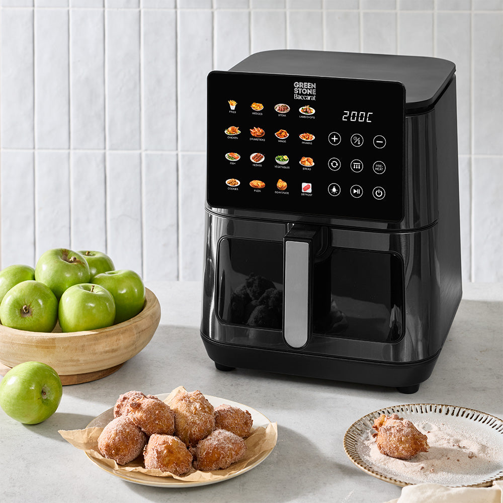 A black digital air fryer is resting on a kitchen countertop alongside green apples in a bowl and fried pastries against a tiled wall backdrop.