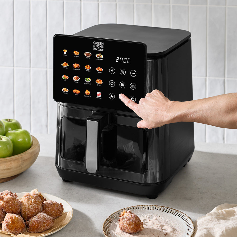 A person's hand is touching the digital touchscreen panel of a black air fryer set on a kitchen counter with snacks and fruit in the background.