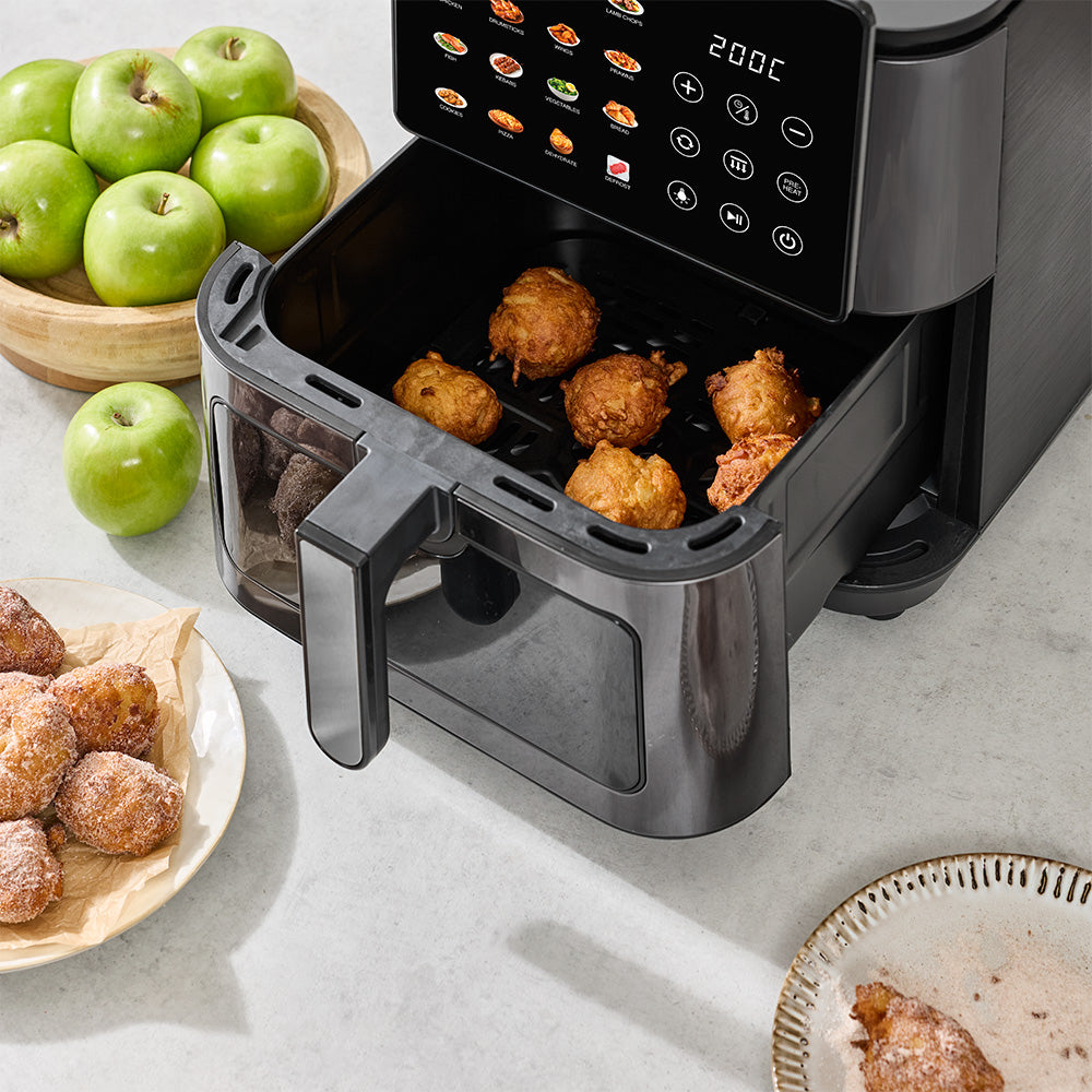A black air fryer sits open on a light countertop, with golden fried balls inside. Green apples are visible in a wooden bowl and on the counter, and plates of fried pastries appear at the edges.