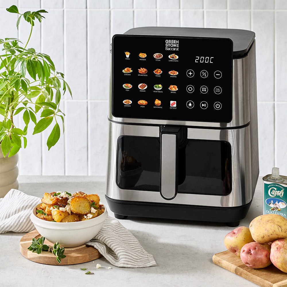 A countertop air fryer in a kitchen setting with a bowl of fried potatoes, whole potatoes, and a potted plant nearby. The air fryer has a black and stainless steel body with a digital touch control screen showing various food options.