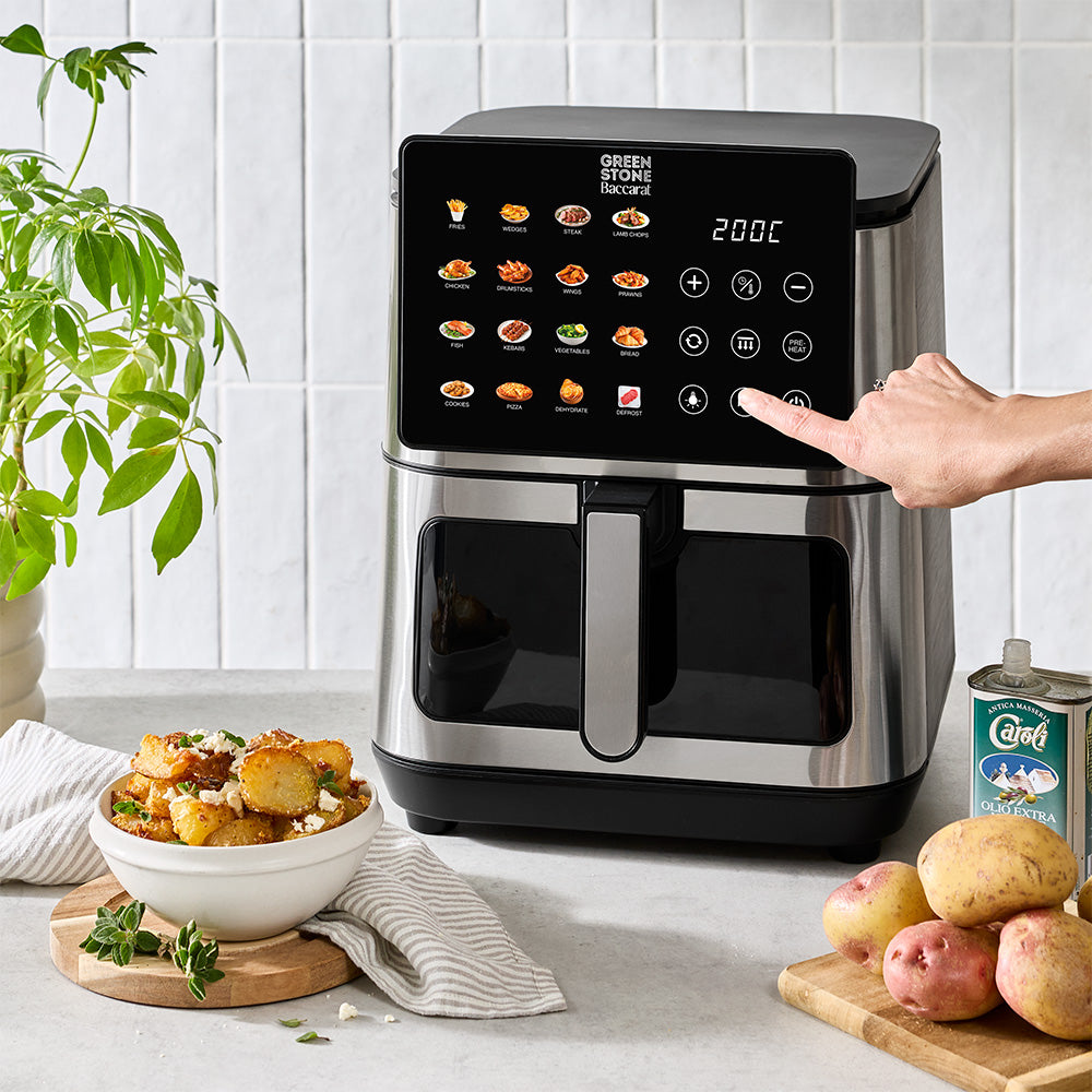 A countertop air fryer in black and stainless steel sits in a kitchen setting with a hand touching its touchscreen interface. A bowl of roasted potatoes, loose potatoes, and a can of green beans are arranged around it on a light stone surface against a white tile backsplash.