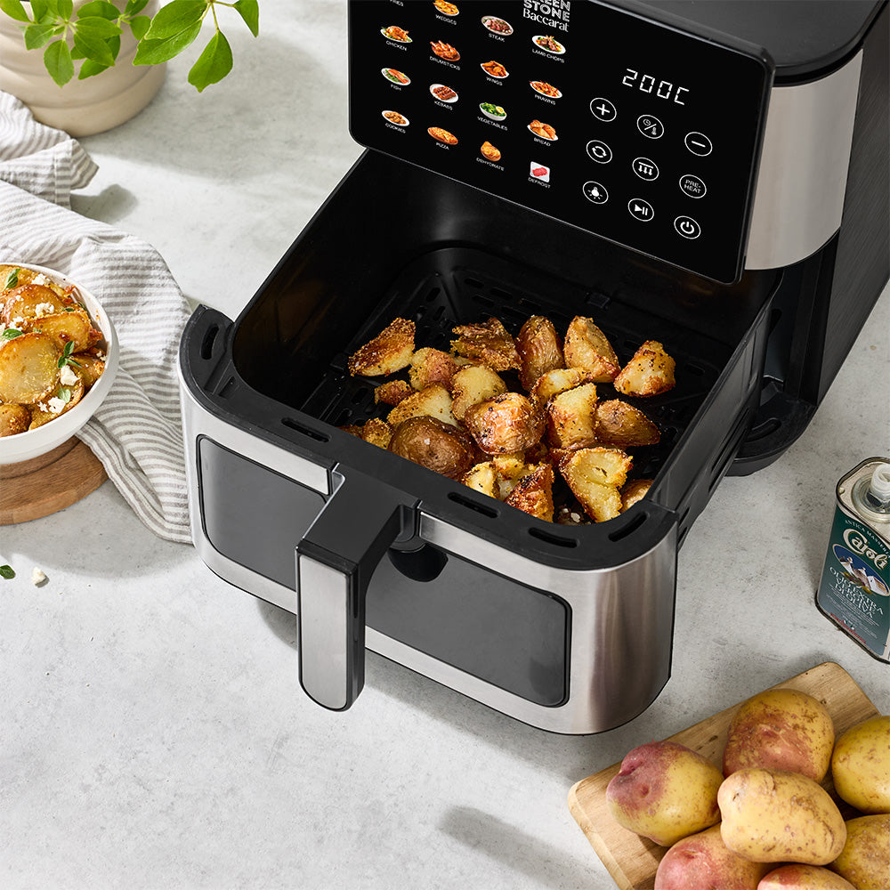 A countertop air fryer with its basket open, filled with roasted potato chunks, in a kitchen setting. A bowl of food, potatoes on a cutting board, and a can of Ford Crownfuls are visible around it.