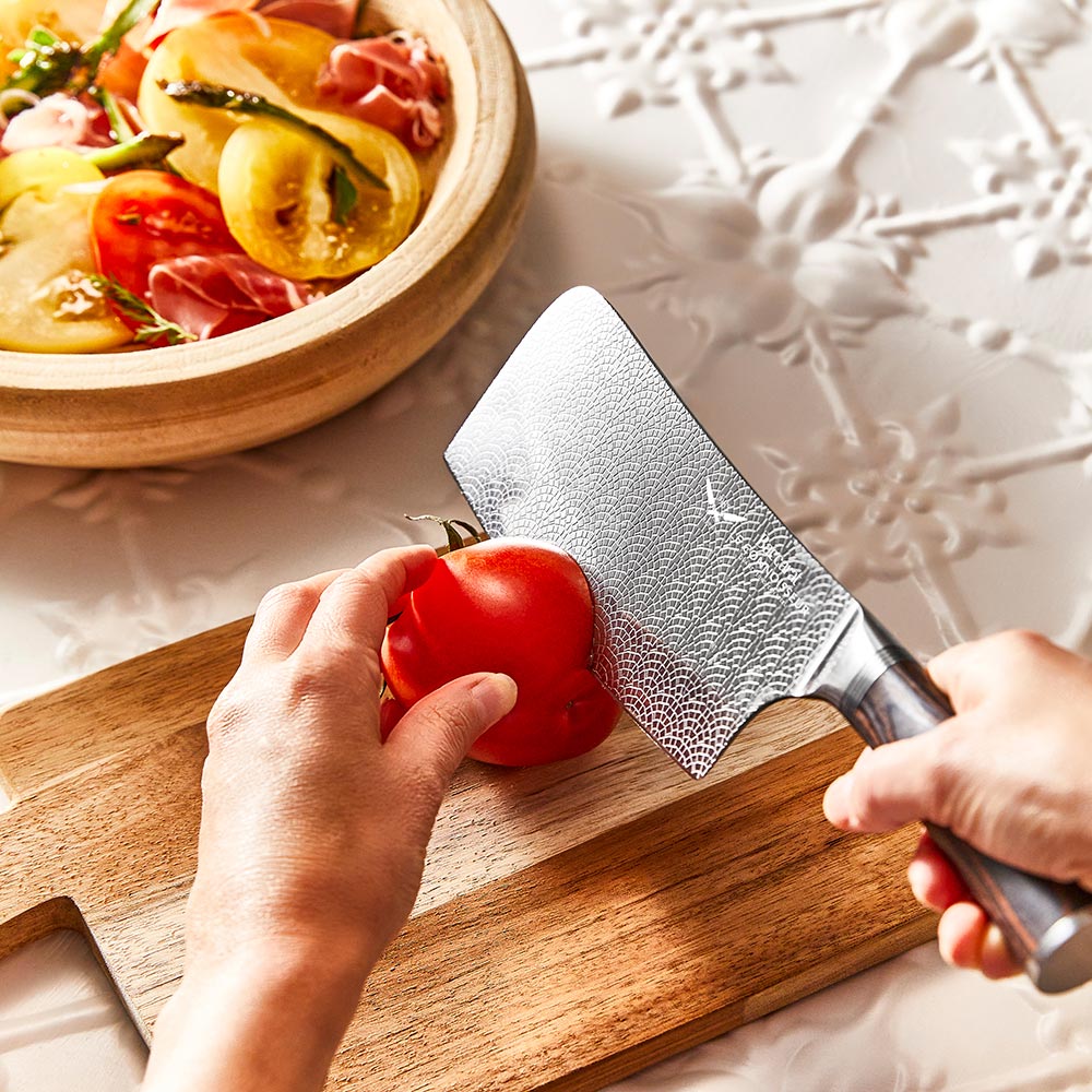 A hand slicing a red tomato with silver cleaver on a wooden cutting board  - Baccarat Le Connoisseur