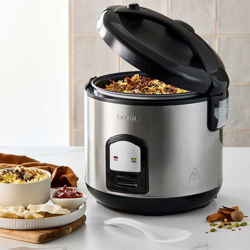 A person opening a silver rice cooker on a kitchen counter next to a bowl of veggies- Baccarat
